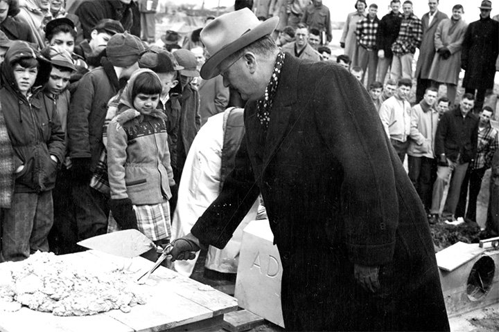A man in an overcoat and hat uses a trowel to spread concrete in a black-and-white photograph.