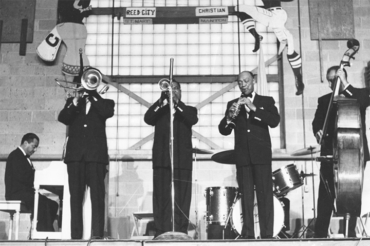Four suit-wearing musicians, one playing an upright base and one playing a trumpet, perform on a stage in a black-and-white photograph.