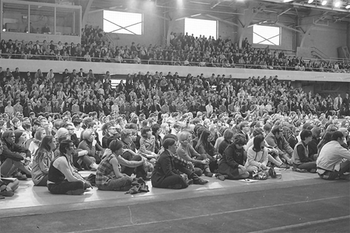 Many people sit on a gymnasium floor and in stadium-style seating facing the right side of a black-and-white photograph.