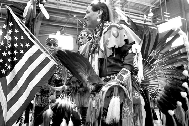 A man dressed in traditional indigenous garb holds an American flag in a black-and-white photo with two other men in the background.