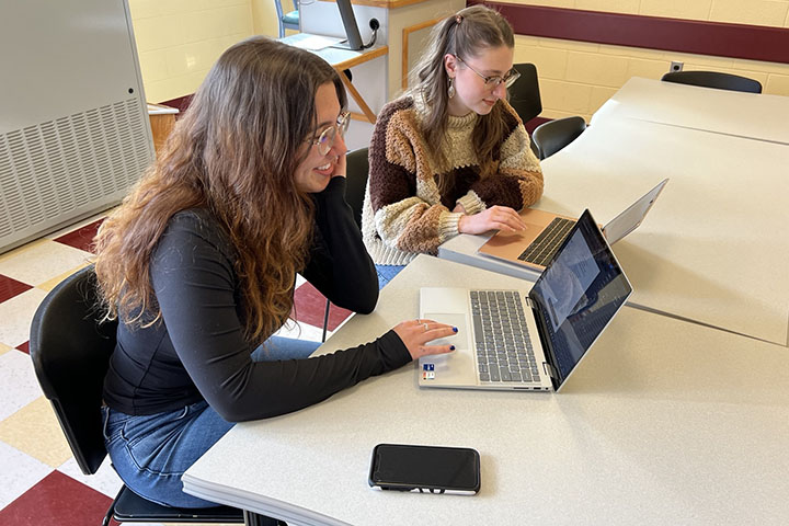 Two young women with long hair and glasses work at laptops on a flat, white table.