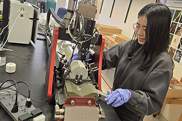 A woman with long black hair, glasses and blue safety gloves examines a rock core sample in a piece of lab equipment.