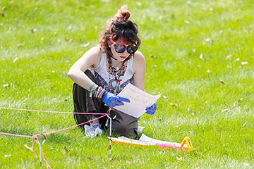 A young woman with a headband and sunglasses looks at a piece of paper in the middle of the grass on a sunny day.
