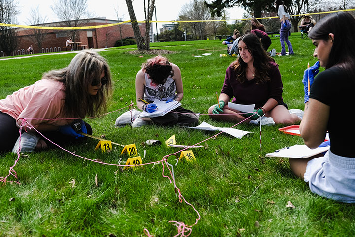 Four young people write on sheets of paper while sitting next to yellow exhibit markers in the grass.