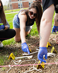 A young woman wearing sunglasses and blue safety gloves points to a fake bone in the grass.