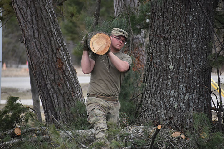A young man in camouflage pants, a camouflage cadet cap and an olive green T-shirt carries a section of tree.