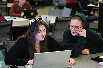 Two young women sit at a table, looking at a laptop computer screen together.