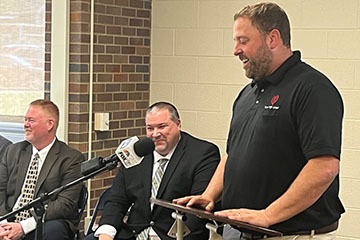 A man in a dark polo shirt speaks from a lectern in front of two other men in suits.