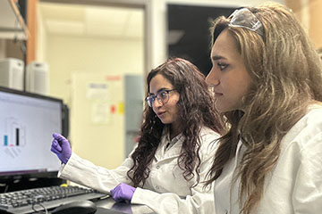 Two young women in white lab coats work at a computer together.