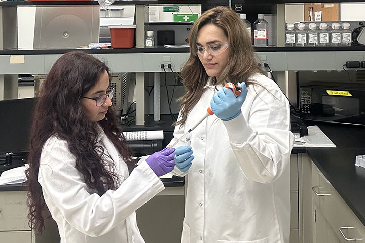 Two long-haired women in safety goggles and lab coats work together on lab samples.