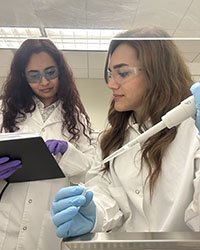 Two long-haired women in safety goggles and lab coats work together on lab samples.