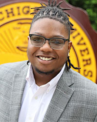 A head shot of a young man wearing a gray blazer and white collared shirt with part of a seal behind him.