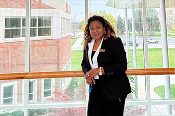 A woman with mid-length curly hair and wearing a black and white suit leans against a railing with a glass wall overlooking a grassy field in the background.