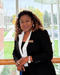 A woman with mid-length curly hair and wearing a black and white suit leans against a railing with a glass wall overlooking a grassy field in the background.