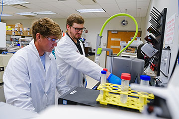 A bearded man with glasses and a white lab coat and blue gloves points at a piece of lab equipment for a young man in safety goggles and a white lab coat.