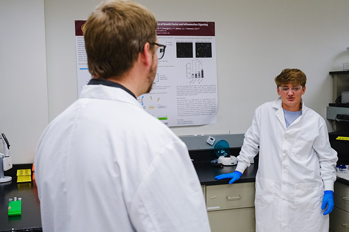 A young man wearing safety goggles, a lab coat and blue safety gloves talks to another man in glasses and a white lab coat.