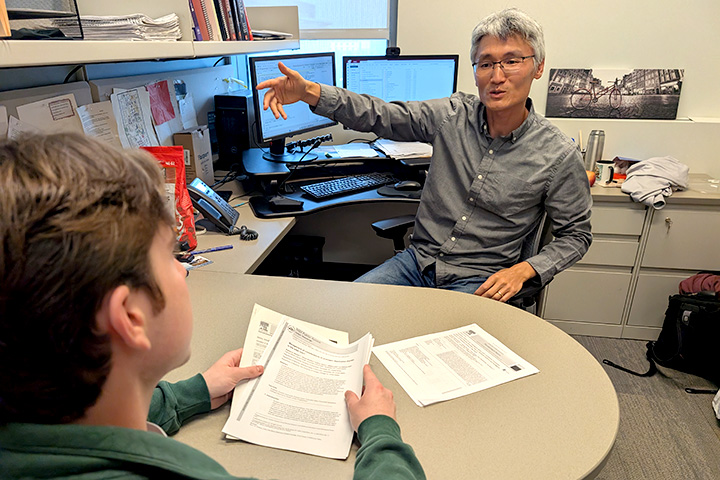 A man gestures while talking to a young man in a green fleece pullover.