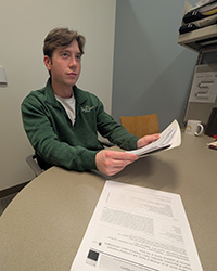 A young man in a green fleece pullover and white T-shirt looks up while holding several pieces of paper.