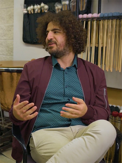 A man with shaggy brown hair wearing a wine-colored jacket and a blue shirt gestures while talking.