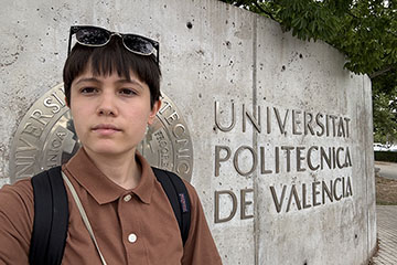 A woman with black hair and sunglasses atop her head stands in from of a stone wall with Universitat Politecnica de Valencia written on it.