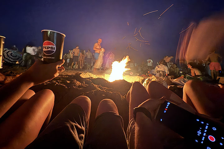 A bonfire burns on a beach with the legs of four people sitting next to it visible in the foreground.