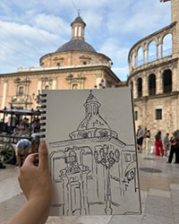 A hand holds a sketch pad with a drawing of an old church with the church in the background.