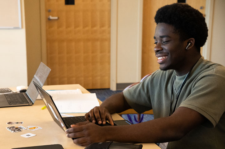 A young man smiles while looking at the screen of his laptop.