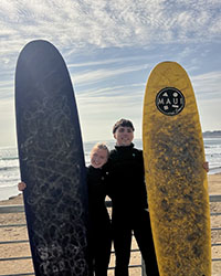 A young man and a young woman in black body suits stand next to each other in between surfboards.