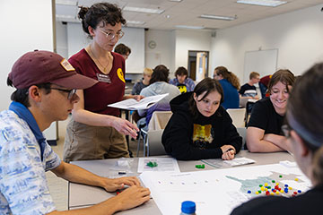 A woman wearing glasses and with black curly hair pulled back into a bun holds a packet of papers in one hand and points at a gaming map with the other while three people sitting at a table look on.