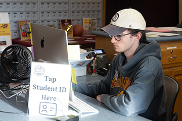 A young man in glasses and a baseball cap looks at a laptop screen with a sign saying, Tap Student ID Here in front.