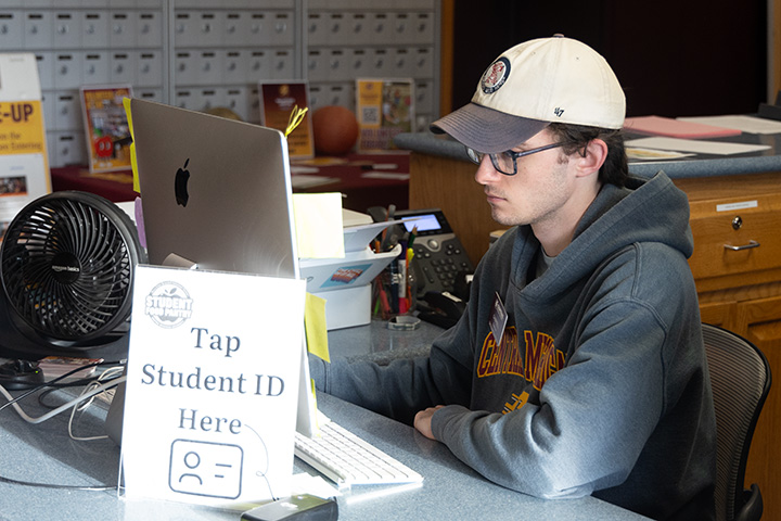 A young man in glasses and a baseball cap looks at a laptop screen with a sign saying, 