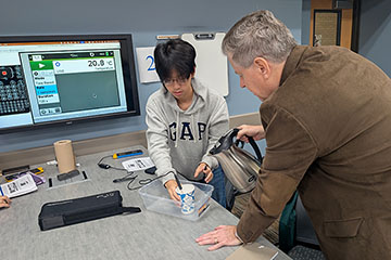 A man with gray hair and wearing a brown jacket pours water into a cup while a young man with glasses holds a temperature probe in it.