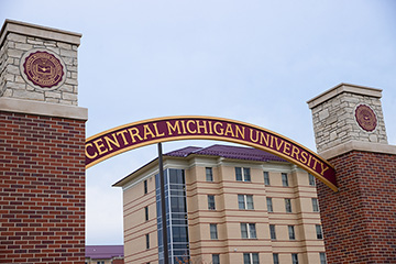 An archway between two brick pillars reads Central Michigan University. A light brown building stands tall in the background.