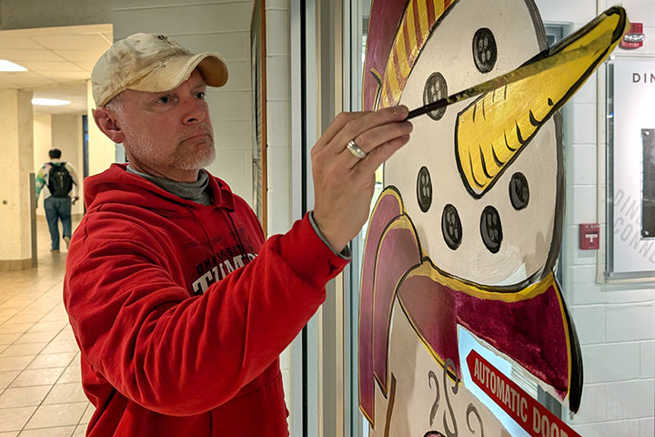 A man in a hat and red sweatshirt paints a cartoon snowman on an indoor window