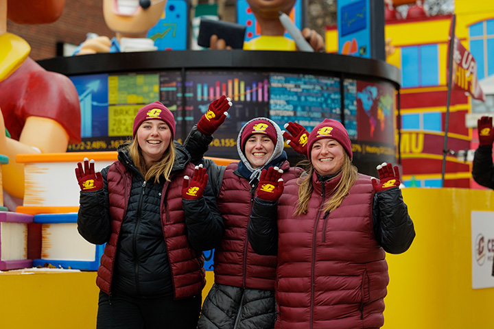 Three people stand in front of the CMU parade float, smiling and waving at the camera
