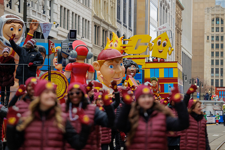The CMU parade float, featuring cartoon-like fire and up-arrow characters, makes its way down through Downtown Detroit