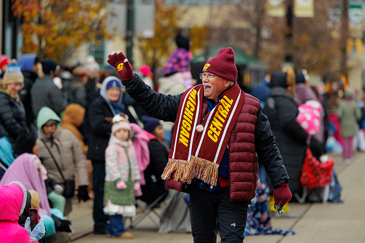 A man with glasses and a gray mustache walks in a parade waving to the crowd while wearing CMU-branded clothing