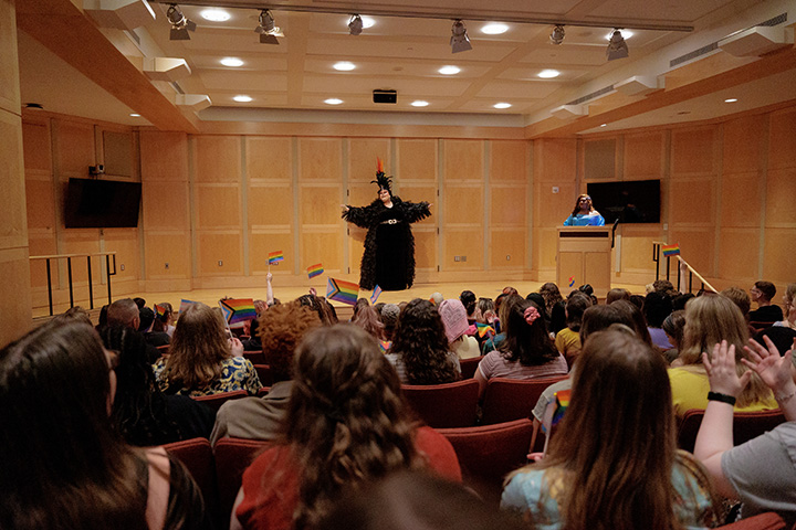 A student dressed in drag stands on a stage in a small auditorium.