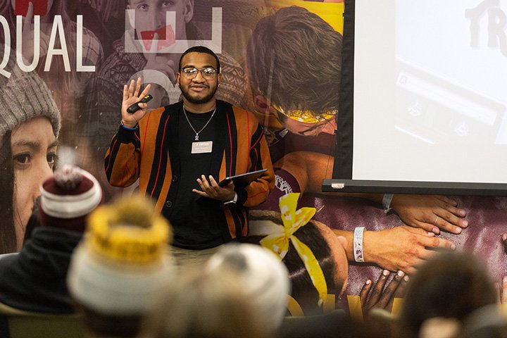 A student stands in front of a crowd waving towards the camera.