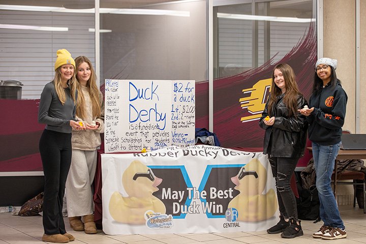 Four female students stand next to a table that has a banner reads May the Best Duck Win written on it.