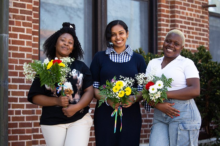 Three female students stand outside a brick building holding flowers.