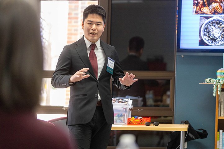 A college student in a suite stands in front of a group giving a presentation.