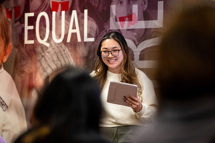 A college student wearing glasses and holding an iPad stands in front of a group of students.