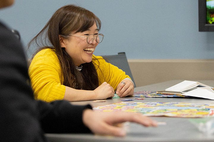 A woman wearing glasses and a yellow long-sleeve shirt sits at a table playing a board game.