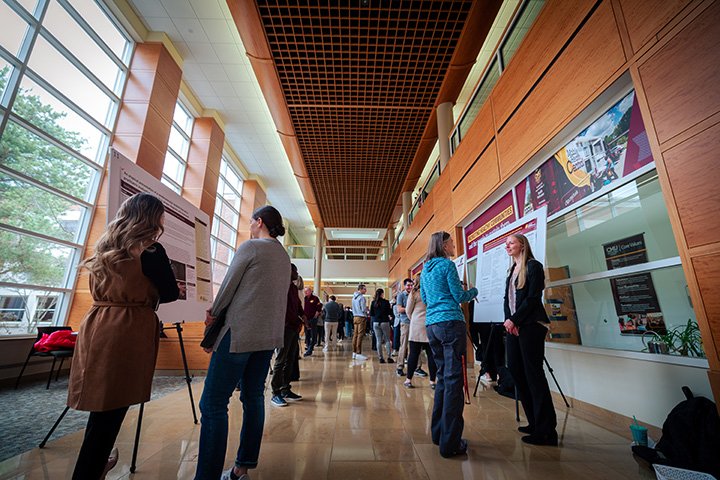 A low, wide-angle shot down a hallway as students stand next to easels with poster boards on display.