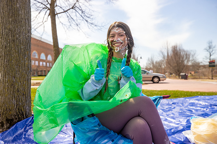 A female college student smiles while having pie smeared all over her face.