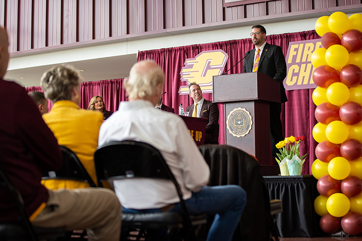 A low angle view of the press conference introducing CMU's new men's basketball coach. Maroon and Gold balloons flank the stage.