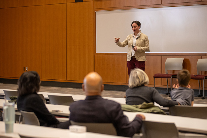 A woman wearing a light brown jacket and white collared shirt talks in front of a classroom.
