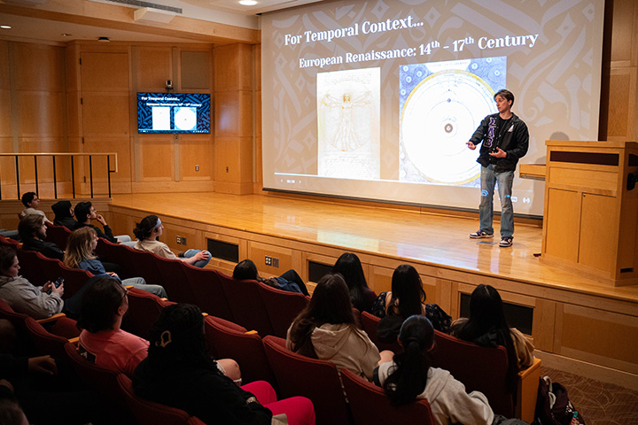 A student stands on stage in a small auditorium giving a presentation about technology.