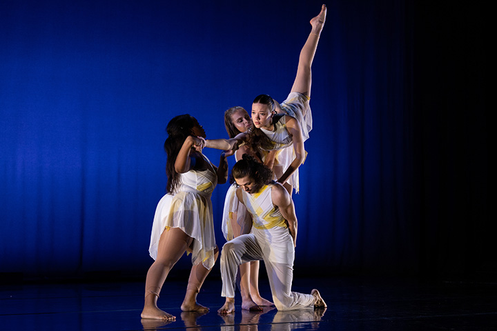 A group of four dancers in white flowing costumes pose on stage. One dancer is lifted up with her leg pointing skyward.
